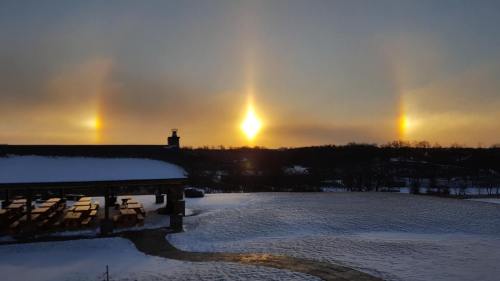 A winter landscape featuring three bright sun halos in a cloudy sky over a snowy field and a building.