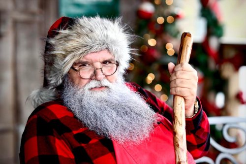 A bearded man in a Santa outfit with glasses and a fur-trimmed hat, holding a wooden staff, surrounded by festive decor.