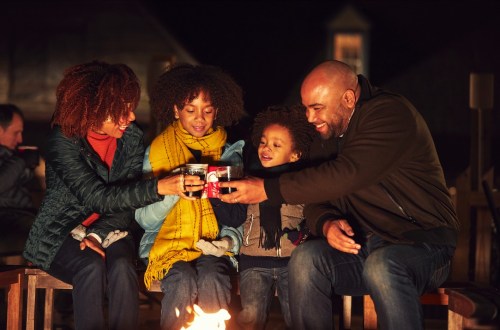 A family gathers around a fire, toasting mugs and enjoying a cozy evening together.