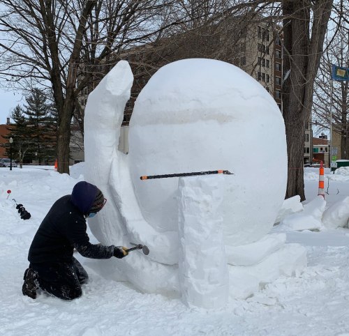 A person chisels a large snow sculpture in a snowy park, with trees and buildings in the background.