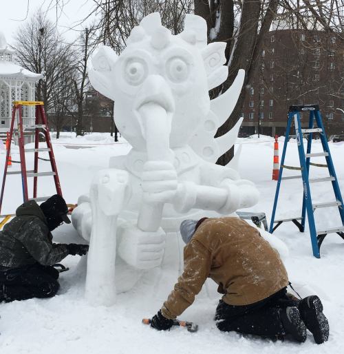Two artists sculpt a large snow figure in a snowy park, surrounded by ladders and trees.
