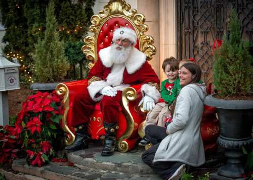 A smiling Santa sits on a throne with a child and a woman, surrounded by festive decorations and plants.