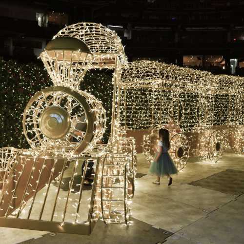 A festive train made of lights, with a child in a green skirt walking beside it in a decorated venue.