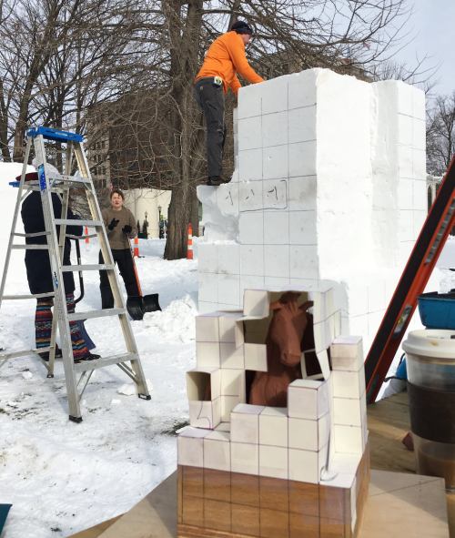 A person on a ladder carves a large snow block, while another person works on a smaller snow sculpture nearby.