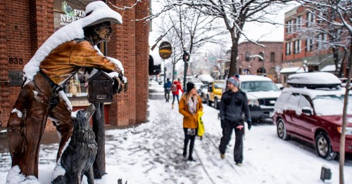 A snowy street scene featuring a cowboy statue, pedestrians, and snow-covered cars in a charming town.