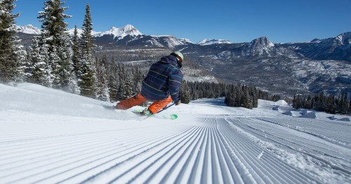 A skier carves through freshly groomed snow on a mountain slope, surrounded by trees and snow-capped peaks.