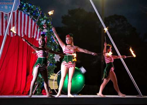 Three performers in festive costumes twirl fire batons on stage, with a holiday-themed backdrop.