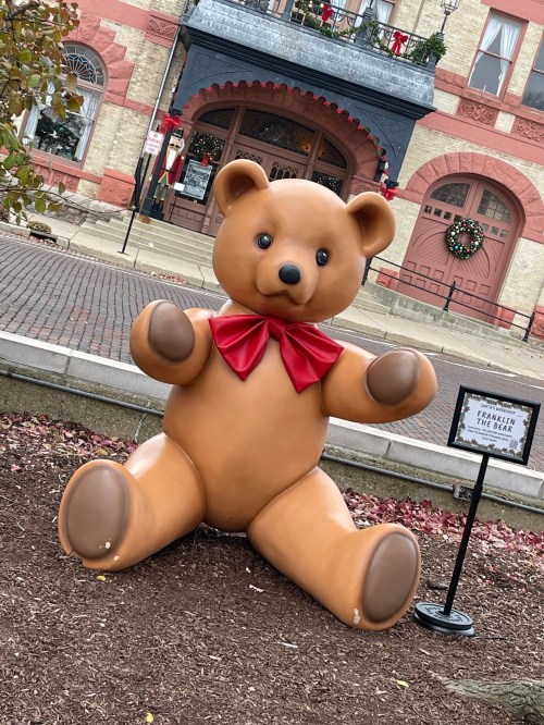 A large, cheerful teddy bear with a red bow sits on the ground in a festive outdoor setting.