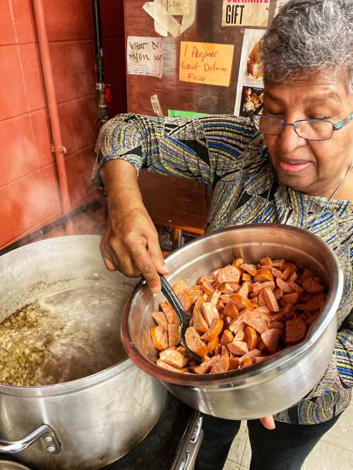A woman stirs a pot of boiling food while adding chopped sausage from a bowl. Steam rises in a kitchen setting.