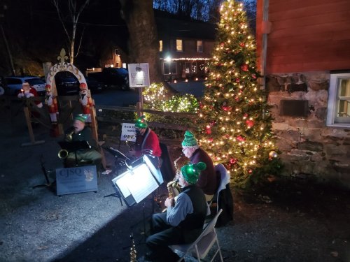 A festive outdoor scene with musicians in green hats playing near a decorated Christmas tree and holiday lights.