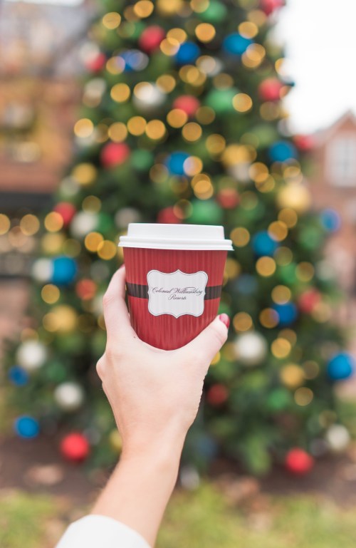 A hand holds a red coffee cup in front of a blurred Christmas tree decorated with colorful ornaments.