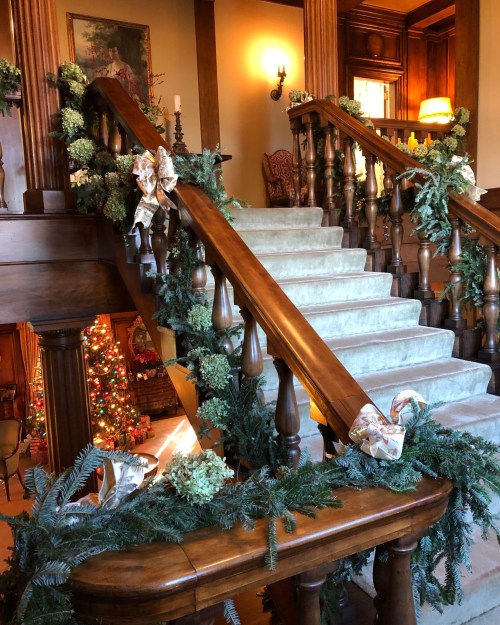 A beautifully decorated staircase with greenery and bows, leading to a cozy room with a Christmas tree in the background.