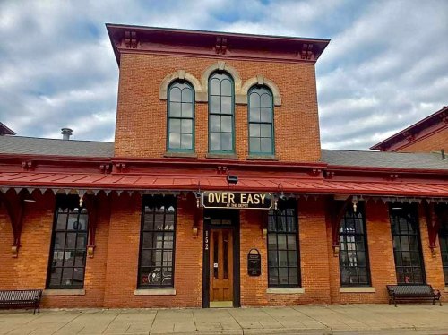 Historic brick building with large windows and a sign reading "Over Easy" above the entrance. Cloudy sky in the background.