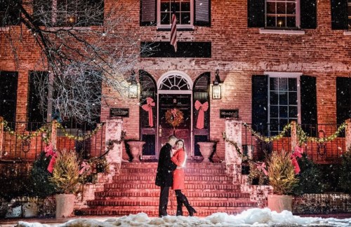 A couple embraces on snowy steps of a festive brick building adorned with holiday decorations and lights.