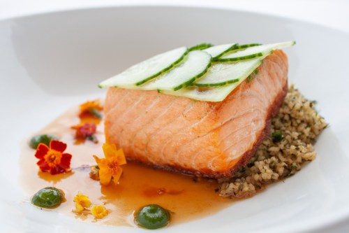 A beautifully plated salmon fillet topped with cucumber slices, garnished with edible flowers and sauce on a bed of grains.