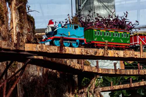 A toy train with a blue engine and green carriages travels along a wooden track in a lush, green setting.