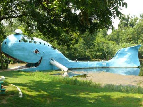 A large blue whale-shaped structure with a slide, situated by a pond and surrounded by greenery.