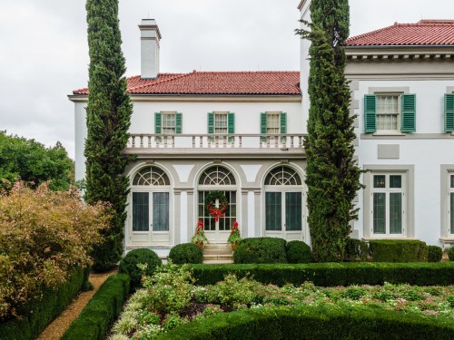 A stately white mansion with green shutters, surrounded by neatly trimmed hedges and colorful flowers.
