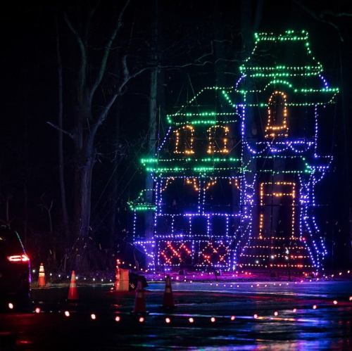 A brightly lit house made of colorful lights, set against a dark background with traffic cones nearby.