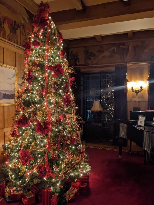 A beautifully decorated Christmas tree with red ornaments and lights, set in a cozy room with a piano and warm lighting.