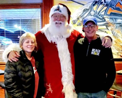 A smiling Santa stands between two adults, one woman and one man, in a festive setting with a colorful background.