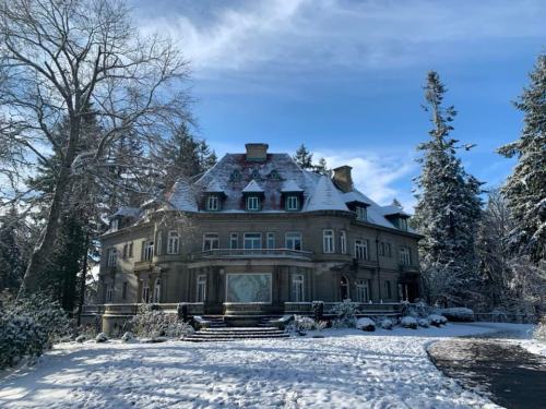 A large, historic mansion covered in snow, surrounded by tall trees under a clear blue sky.