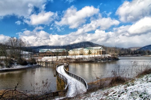 A winding boardwalk leads to a snowy lakeside with mountains and a cloudy blue sky in the background.