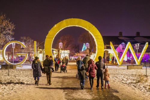 A winter scene with people walking under a large illuminated "GLOW" sign, surrounded by festive lights and snow.