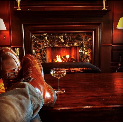 Cozy scene with brown boots resting on a table, a glass of drink, and a warm fireplace in a stylish room.