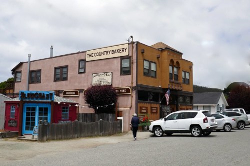 A quaint bakery building with a sign reading "The Country Bakery," surrounded by shops and parked cars on a cloudy day.