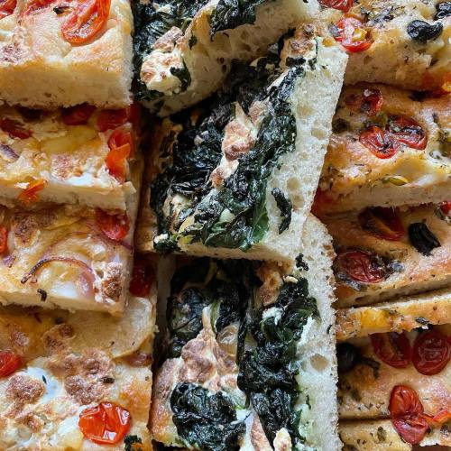 A close-up of assorted focaccia bread topped with tomatoes, spinach, and cheese, arranged in a colorful display.