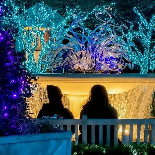 Two people sit on a bench in front of a glowing fountain, surrounded by blue and white lights.