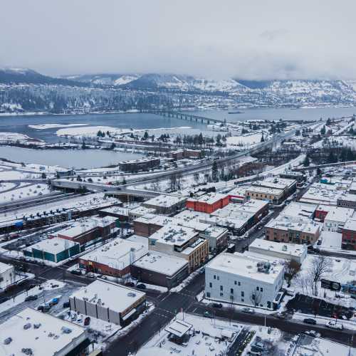 Aerial view of a snowy town with rooftops covered in snow, a river, and mountains in the background.