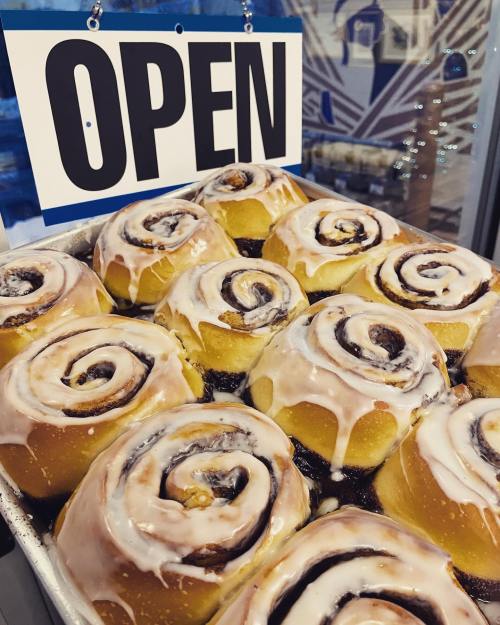 Freshly baked cinnamon rolls with icing, displayed in a tray, next to an "OPEN" sign.