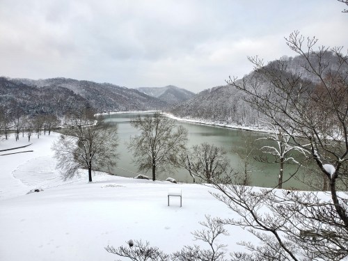 A serene winter landscape featuring a snow-covered river surrounded by mountains and bare trees.