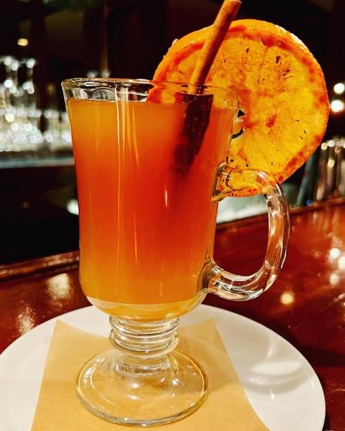 A warm drink in a glass mug, garnished with a cinnamon stick and a slice of caramelized orange on the rim.