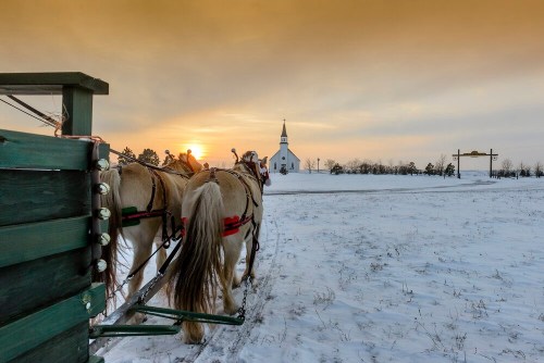 Horses pulling a sled in snow, with a church and sunset in the background.