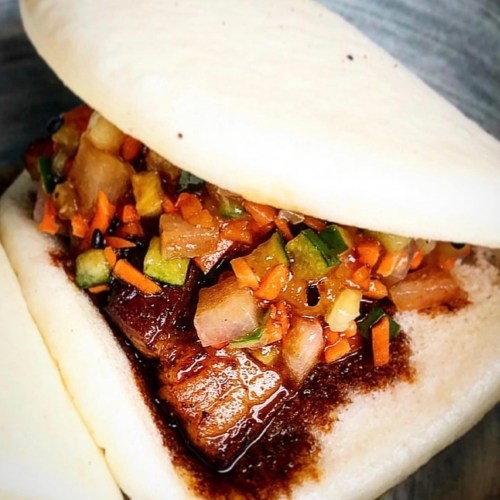 A close-up of a bao bun filled with marinated meat and a colorful vegetable salsa.