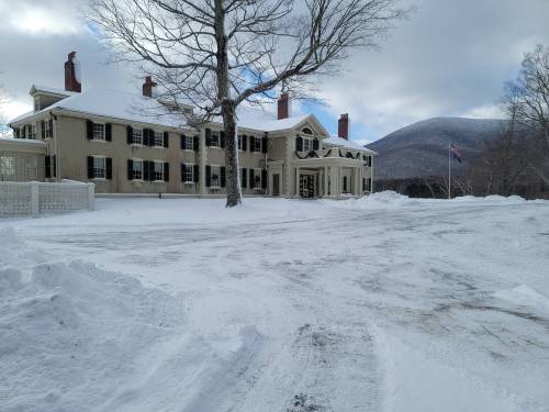 A large, historic house covered in snow, with a tree in front and mountains in the background under a cloudy sky.