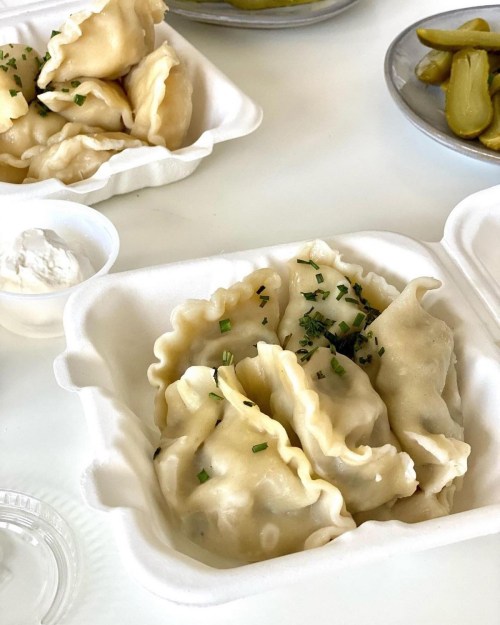 A takeout container filled with dumplings, garnished with chives, alongside pickles and a small cup of sour cream.