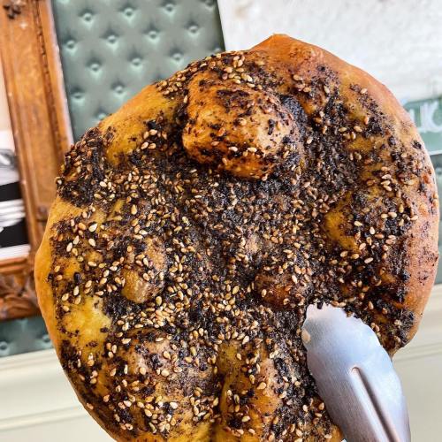 A round, golden-brown bread topped with sesame seeds and a dark, textured crust, with a serving utensil beside it.