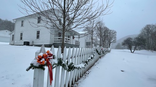 A snowy scene featuring a white house and a picket fence adorned with holiday decorations. Trees are bare in the background.