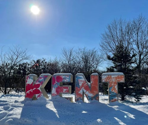 Colorful "KENT" sign surrounded by snow under a bright blue sky and sun. Trees in the background.