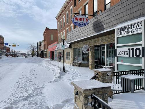 Snow-covered street with shops and a sign for downtown living, showcasing a quiet winter scene.