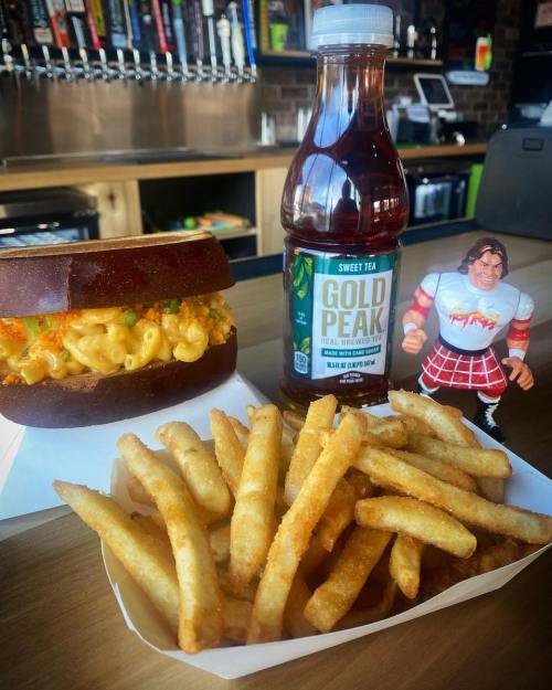 A sandwich with macaroni, a bottle of sweet tea, and a serving of fries next to a toy figure on a table.