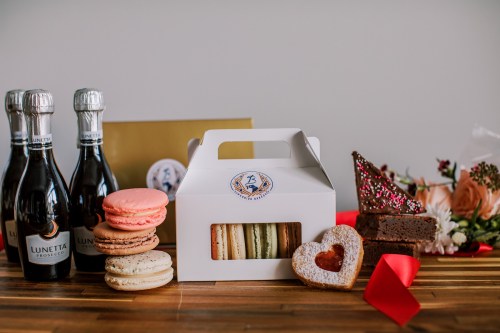 A display of macarons, a heart-shaped cookie, brownies, sparkling wine, and a bouquet on a wooden surface.