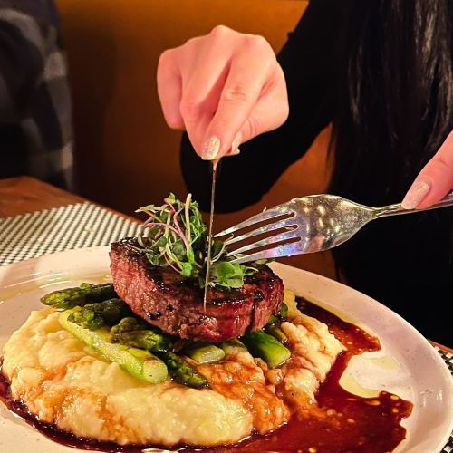 A hand holds a fork and knife, cutting into a steak atop creamy mashed potatoes and asparagus, garnished with microgreens.