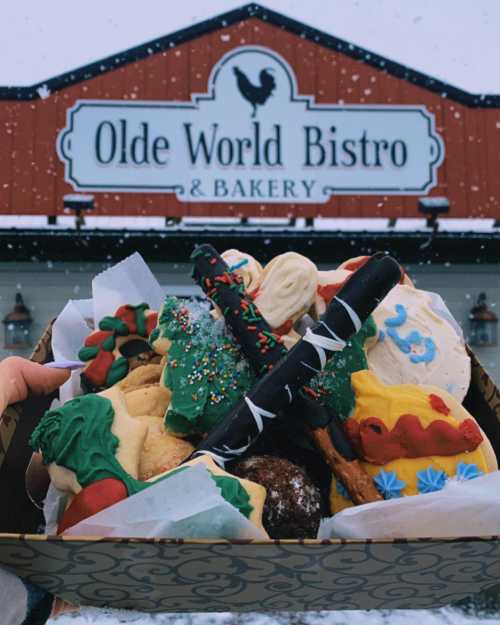 A hand holds a box of colorful holiday cookies in front of Olde World Bistro & Bakery, with snow falling around.