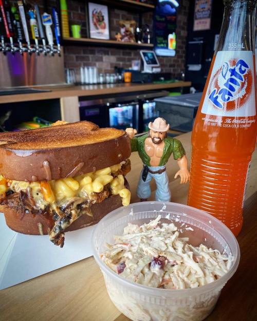 A grilled sandwich filled with mac and cheese, a bottle of orange soda, coleslaw, and a toy figure on a table.