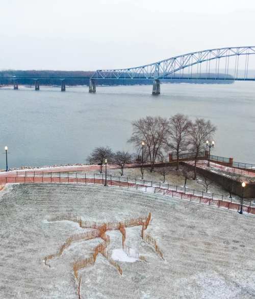 A snowy landscape with a large spider sculpture in the foreground and a bridge over a river in the background.